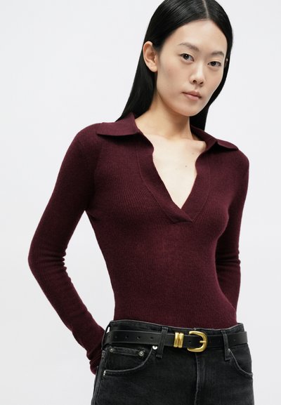 Νεαρή γυναίκα με μακριά μαύρα μαλλιά φορά εφαρμοστό βυσσινί πουλόβερ με V λαιμόκοψη και μαύρο τζιν με μαύρη ζώνη που έχει χρυσή αγκράφα.