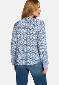 Blouse blanche avec motif abstrait bleu, manches longues, empiècement plissé au dos, associée à un jean foncé, avec un ourlet arrondi.