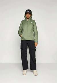 Žmogus, vilkintis žalią striukę su kapišonu, juodas krovinių kelnes, šviesiai rudas lauko batus ir juodą kepurę, stovi priešais vientisą šviesų foną.