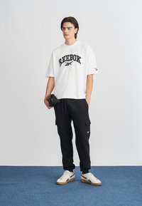 Bijela oversize majica s crnim "Reebok" logom, uparena s crnim cargo trenirkama i bijelim tenisicama s crnim akcentima.
