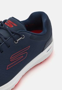 Navy athletic shoe with a textured synthetic upper, red accents, and a white sole. Features include lace-up design and padded collar.