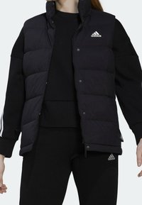 Svart isolerad puffer-väst med hög krage och tryckknappar, bärs över en svart sweatshirt. Har en vit Adidas-logga på bröstet.