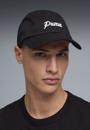 Joven con una gorra de béisbol negra de Puma y camiseta negra, mirando ligeramente hacia un lado sobre un fondo gris liso.