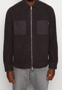 Giacca zip-up grigio scuro con colletto, dotata di due tasche sul petto e una texture liscia. I polsini elasticizzati e il dettaglio dell'orlo migliorano la vestibilità.