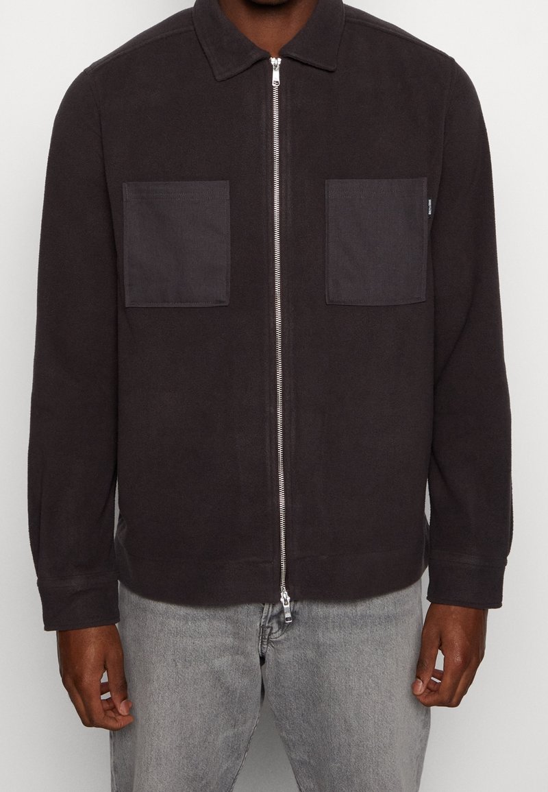 Giacca zip-up grigio scuro con colletto, dotata di due tasche sul petto e una texture liscia. I polsini elasticizzati e il dettaglio dell'orlo migliorano la vestibilità.