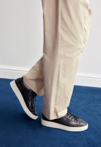 Črni usnjeni supergi z belo gumijasto podplatom, s gladko teksturo in klasičnim vezalkami, kombinirani z bež hlačami na modrem preprogi.