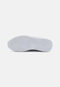 Semelle d'une sneaker en caoutchouc blanc, avec un motif en chevrons et un logo rectangulaire au centre.