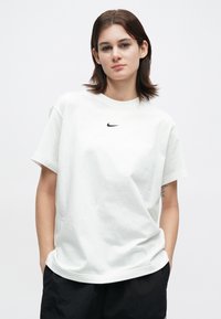 Fiatal személy, rövid barna hajjal, fehér Nike pólót és fekete sortot viselve, a zsebeit a zsebében tartva áll, egy sima fehér háttér előtt.