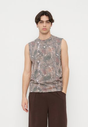 Giovane uomo con capelli castano scuro di media lunghezza, che indossa una maglietta senza maniche con motivo mimetico e pantaloni marrone scuro, in piedi davanti a uno sfondo bianco.