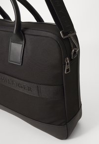Schwarze Stofftasche mit strukturierter Oberfläche, ausgestattet mit einem stabilen oberen Griff, verstellbarem Riemen und einem auffälligen Logo-Band. Reißverschluss-Detail.