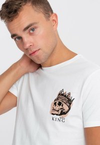 Bílý bavlněný tričko s grafickým potiskem korunované lebky a slovem "KING" černě umístěným níže. Krátké rukávy, design s kulatým výstřihem.