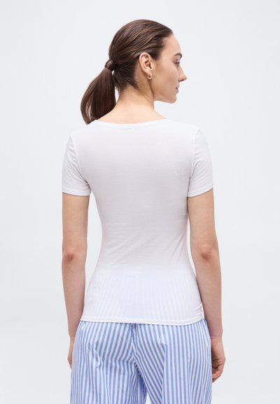 Γυναίκα με καστανά μαλλιά σε αλογοουρά, φορώντας εφαρμοστό λευκό κοντομάνικο πουκάμισο και μπλε-λευκό παντελόνι με κάθετες ρίγες, με την πλάτη γυρισμένη.