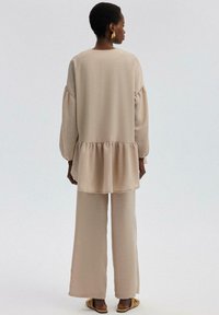 Blusa beige, ligera, con mangas abullonadas y dobladillo acampanado, combinada con pantalones anchos a juego y sandalias planas. Tejido texturizado.