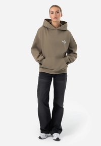 Felpa verde oliva con tasca frontale e logo bianco, abbinata a jeans neri a gamba larga e scarpe sportive grigie. Texture e design sottili.