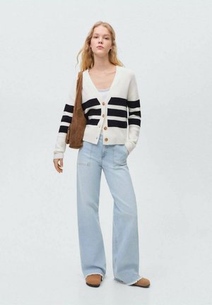 Cardigan alb și bleumarin cu dungi, cu cinci butoane, purtat cu blugi largi de culoare albastru deschis și încălțăminte maro, ținând o geantă din material.