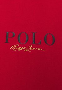 Tissu rouge avec texte noir « POLO » et broderie cursive jaune « Ralph Lauren » sous le texte, centré sur le matériau.