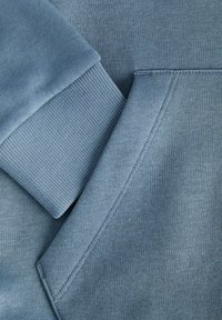 Mėlynas džemperis, kuris demonstruoja tekstūruotą medžiagą, raukšlėtus rankogalius ir kengūros kišenę su tvarkingomis siūlėmis. Lygus paviršius ir švelnus jausmas.