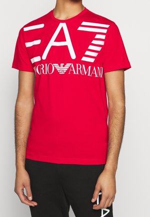 Mężczyzna w jaskrawoczerwonej koszulce EA7 Emporio Armani z dużym białym logo na przodzie oraz czarnych spodniach z częściowym białym napisem.