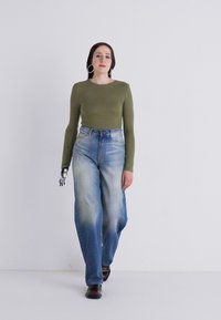 Olivengrønn langermet topp kombinert med høyt liv, lyseblå, falmede jeans. Inkluderer en svart protesthånd og brune sko. Enkelt bakgrunn.