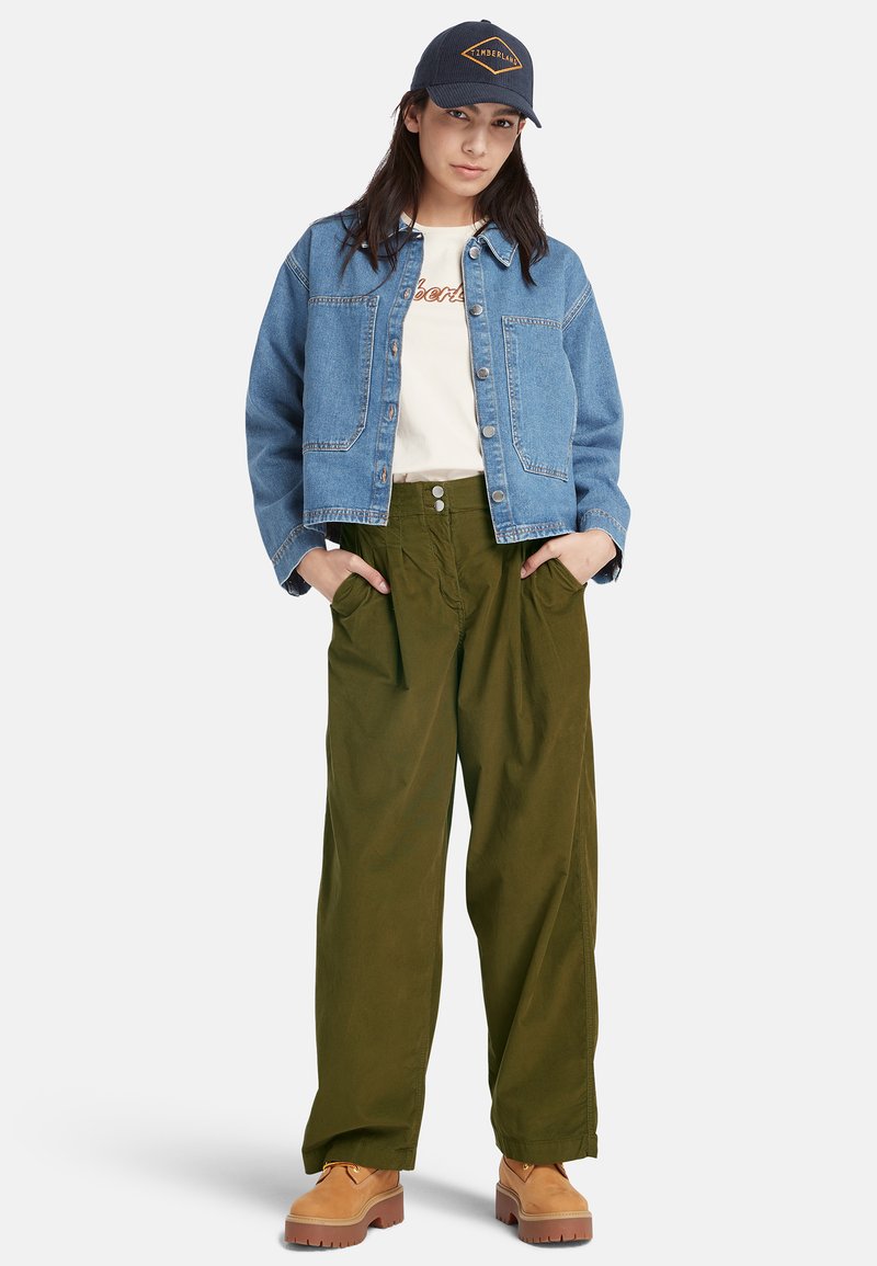 Jeansjacke in Hellblau mit zwei Fronttaschen, getragen über einem weißen T-Shirt. Olivgrüne weite Hosen und braune Schuhe runden den Look ab.