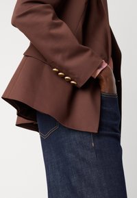 Brun blazer med en glatt tekstur, med en lett utsvingt hem, detaljer med gullknapper på mansjetten, sammen med mørk denim jeans.