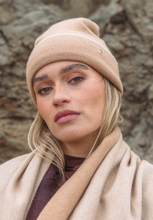 Hellbraune gestrickte Mütze mit einem weißen Streifen und einem kleinen metallischen Akzent. Weiche Textur. Modell trägt einen beigen Schal über einem dunklen Oberteil.