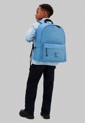 Gutt iført lyseblå skjorte, mørke bukser, svarte sko, med en stor lyseblå Calvin Klein-ryggsekk, stående foran en ensfarget bakgrunn.