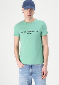 Mint green T-shirt with short sleeves featuring "TOMMY HILFIGER EST. 1985" in navy text. Worn with light blue, slightly distressed jeans.