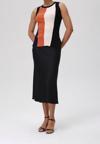 Woman wearing color-block sleeveless top with black, white, orange, and navy panels, black midi skirt, and black ankle-strap sandals.