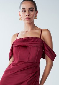 Burgunderfarbenes Satin-Kleid mit schulterfreien Ärmeln, gefaltetem Oberteil und drapierten Details. Goldene Creolen ergänzen den Look.