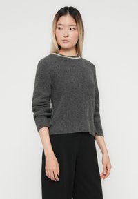 Pull en tricot gris avec un décolleté arrondi, coupe ample et manches retroussées. La texture semble douce et confortable avec un léger motif côtelé.