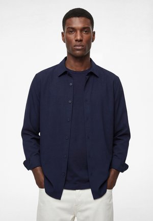 Mann mit kurzen Haaren, der ein navyblaues Hemd über einem dunkelblauen T-Shirt und weiße Hosen trägt und mit den Händen in den Taschen steht.