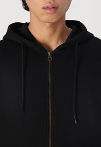 Sudadera con cremallera negra hecha de una tela suave, con una cremallera frontal con herrajes de metal y cordones para la capucha. Textura lisa.