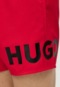 Κόκκινα μαγιό με πλαϊνή τσέπη και μεγάλο μαύρο λογότυπο "HUGO" τυπωμένο στον μηρό πάνω από ανοιχτόχρωμη τρίχα ποδιού.
