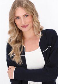 Marineblauer Cardigan mit weißem Besatz und weicher Textur, getragen über einem weißen Oberteil. Lange Ärmel und offener Schnitt schaffen eine entspannte Silhouette.