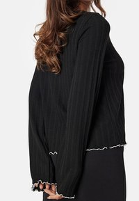 Maglione a coste nero con maniche lunghe, design cropped e dettagli in pizzo bianco su orlo e polsini. Texture morbida con una silhouette attillata.