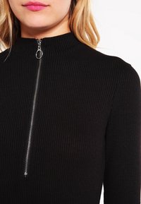 En blond kvinna som bär en svart ribbad tröja med en silverfärgad dragkedja som går vertikalt från kragen till mitten av bröstet.