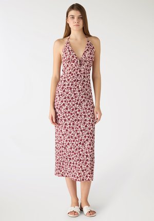 Giovane donna in piedi che indossa un abito midi senza maniche con scollo all'americana, stampa floreale rossa e sandali bianchi modello slide su uno sfondo semplice.