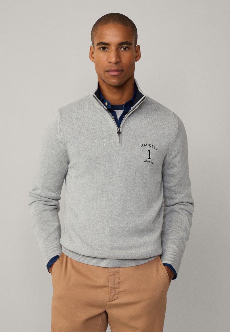 Suéter gris claro con cremallera, con puños y dobladillo acanalados, que presenta un pequeño logo en el lado izquierdo del pecho. Usado sobre una camisa de color azul con cuello.