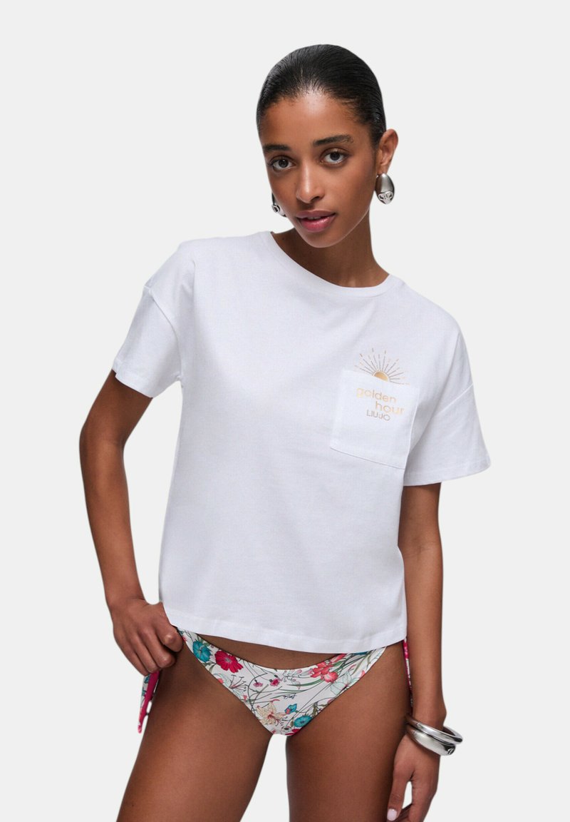 Femme portant un T-shirt blanc avec le texte « golden hour » et un motif de soleil, un bas de bikini à fleurs, des boucles d’oreilles créoles argentées et des bracelets sur un fond uni.