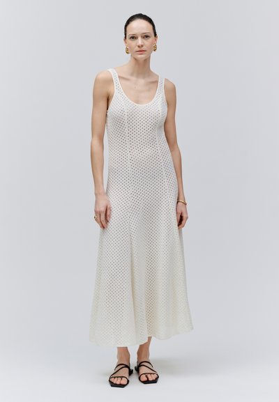 Femme debout portant une robe maxi blanche sans manches en crochet, des sandales noires à brides, des boucles d’oreilles créoles dorées et des bijoux minimalistes.