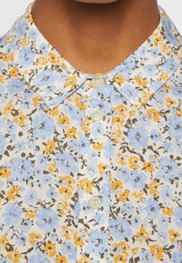 Gros plan sur une chemise à col avec un motif floral bleu, jaune et blanc et trois boutons blancs visibles fermés au niveau du col.