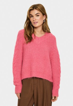 Frau mit hellbraunem Haar, die silberne Ohrringe, einen strukturierten rosa Strickpullover und braune weite Hosen trägt, vor einem schlichten Hintergrund.