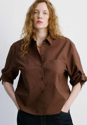 Ung kvinde med langt rødt hår, iført en løs brun skjorte med knapper og opkørte ærmer samt mørke bukser, hænderne i lommerne, neutral baggrund.