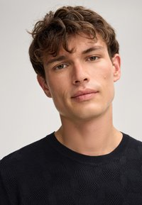 Pullover nero in maglia con texture, caratterizzato da un motivo geometrico, scollatura rotonda e finitura liscia. Il modello ha capelli corti e scompigliati.