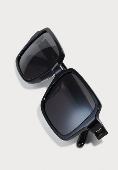 Lunettes de soleil oversize noires en forme de carré avec des verres foncés. Fabriquées en plastique brillant, elles présentent des montures épaisses et un design angulaire distinct.