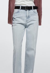 Femme portant un jean droit taille haute bleu clair avec poches avant, passants de ceinture et une ceinture en cuir noire, assorti à un haut blanc rentré.