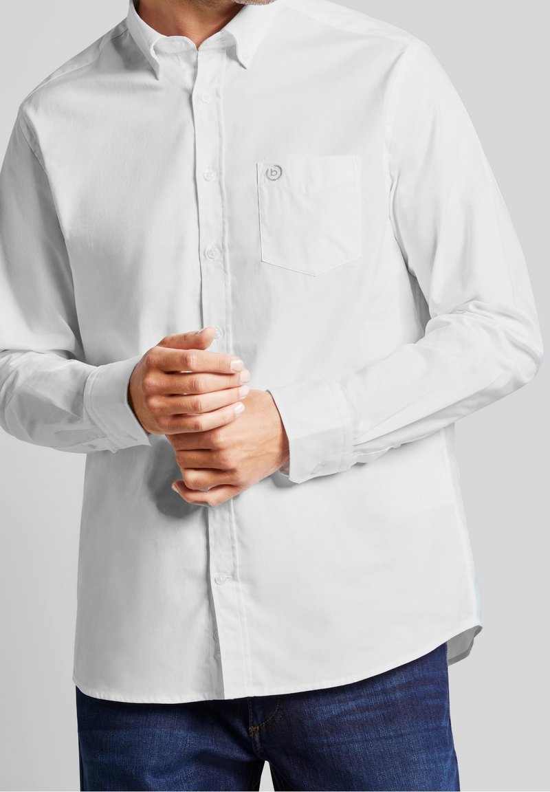 Homme portant une chemise blanche à boutons avec une poche poitrine et un jean bleu, mains jointes devant, visible du cou jusqu'aux hanches.