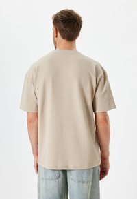 Beige tekstureret kortærmet skjorte med rund halsudskæring. Har et subtilt mønster og en løs pasform, parret med lyseblå denimbukser.