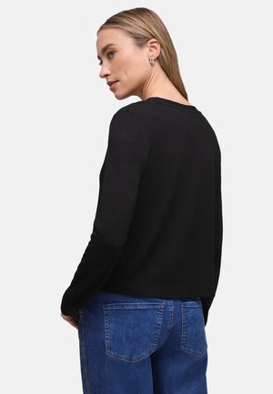 Frau mit blondem Haar, trägt ein schwarzes Langarmshirt und blaue Jeans, von hinten vor einem schlichten Hintergrund gezeigt.